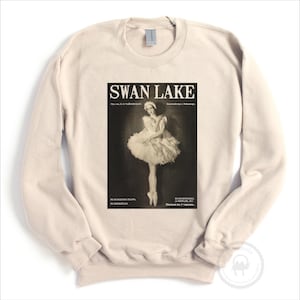 May include: Cream-colored sweatshirt featuring a vintage "Swan Lake" poster design. The poster depicts a ballerina in a tutu, with text in English and Russian. The sweatshirt has long sleeves and a crew neck.