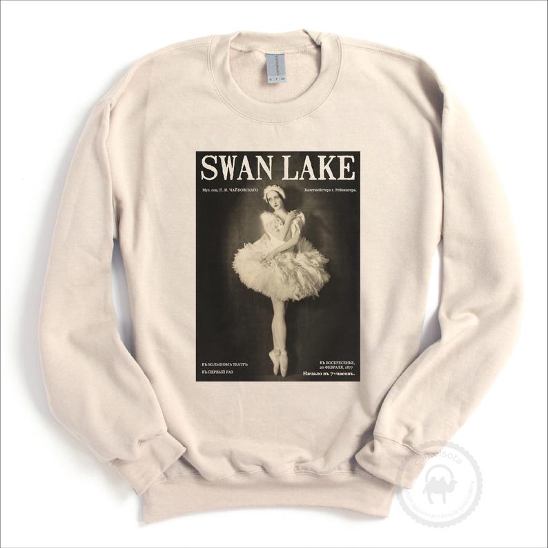 May include: Cream-colored sweatshirt featuring a vintage "Swan Lake" poster design. The poster depicts a ballerina in a tutu, with text in English and Russian. The sweatshirt has long sleeves and a crew neck.