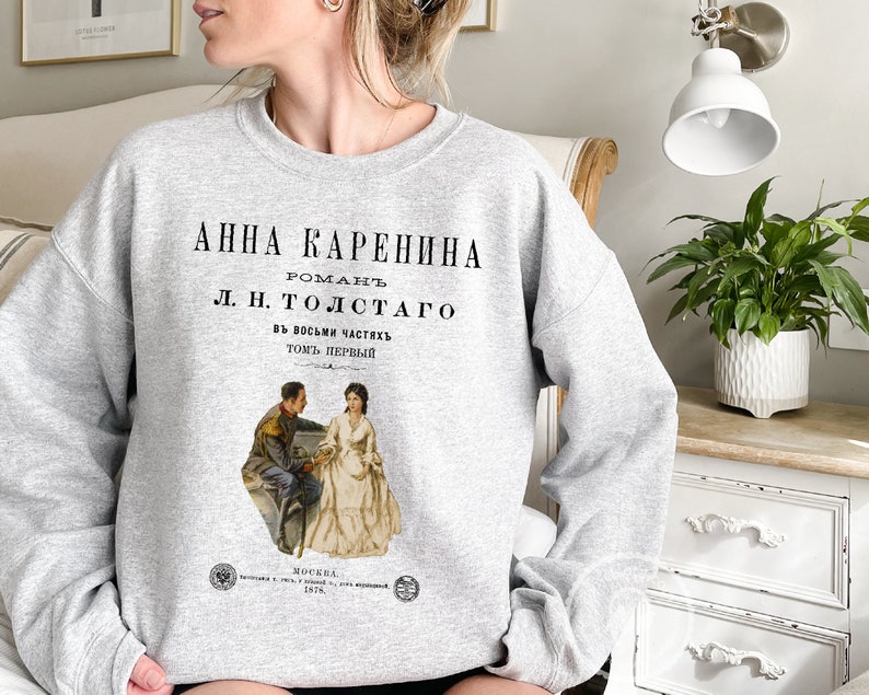 May include: Gray sweatshirt with a vintage book cover design featuring a black and white illustration of a couple. The text on the cover reads "Anna Karenina, Roman L.N. Tolstoy, in eight parts, volume one".