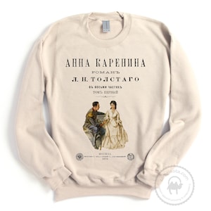 May include: A beige sweatshirt with a vintage-style print of the book cover for "Anna Karenina" by Leo Tolstoy. The cover features an illustration of two people in 19th-century clothing.