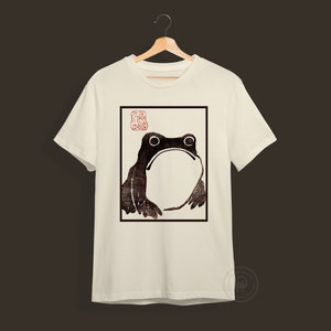 May include: White t-shirt with a black and white image of a frog in a square frame. The image is a woodblock print style. The frog is looking up and has a large belly. There is a red stamp in the top left corner of the frame.