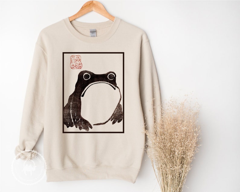 May include: A beige sweatshirt with a black and white illustration of a frog in a square frame. The frame has a red stamp with Japanese characters in the top left corner.