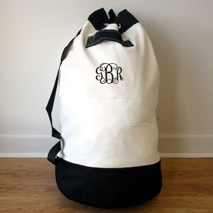 May include: A large, white canvas duffel bag with a black base and accents. The bag features a drawstring closure and black shoulder straps. A black monogram with the letters "BR" is centered on the front.