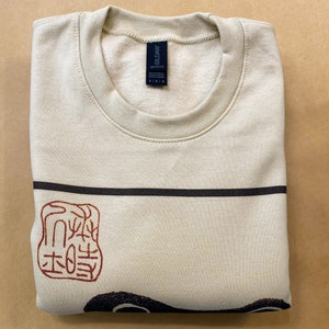 May include: A beige sweatshirt with a black and red graphic print of a Japanese stamp. The stamp features a red design on a white background. The sweatshirt has a crew neck and is made of a soft, comfortable fabric.