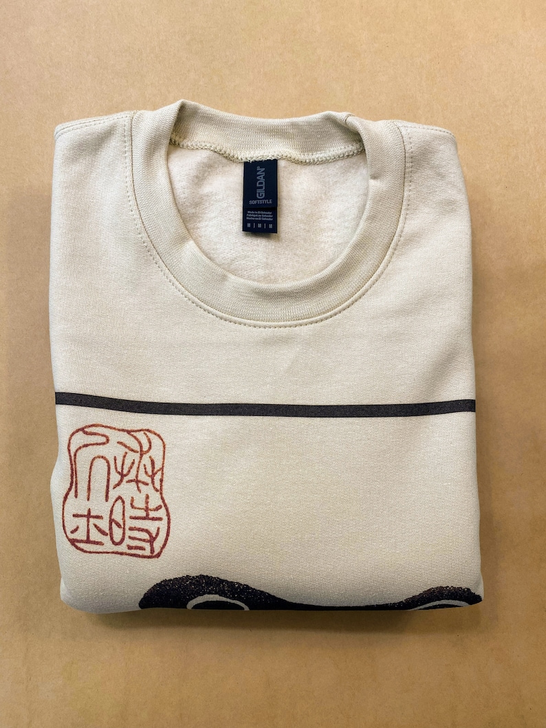 May include: A beige sweatshirt with a black and red graphic print of a Japanese stamp. The stamp features a red design on a white background. The sweatshirt has a crew neck and is made of a soft, comfortable fabric.