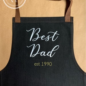 K&ouml;nnte beinhalten: Schwarze Denim-Sch&uuml;rze mit braunen verstellbaren Tr&auml;gern. Die Sch&uuml;rze hat wei&szlig;en Text, der "Best Dad est. 1990" lautet.