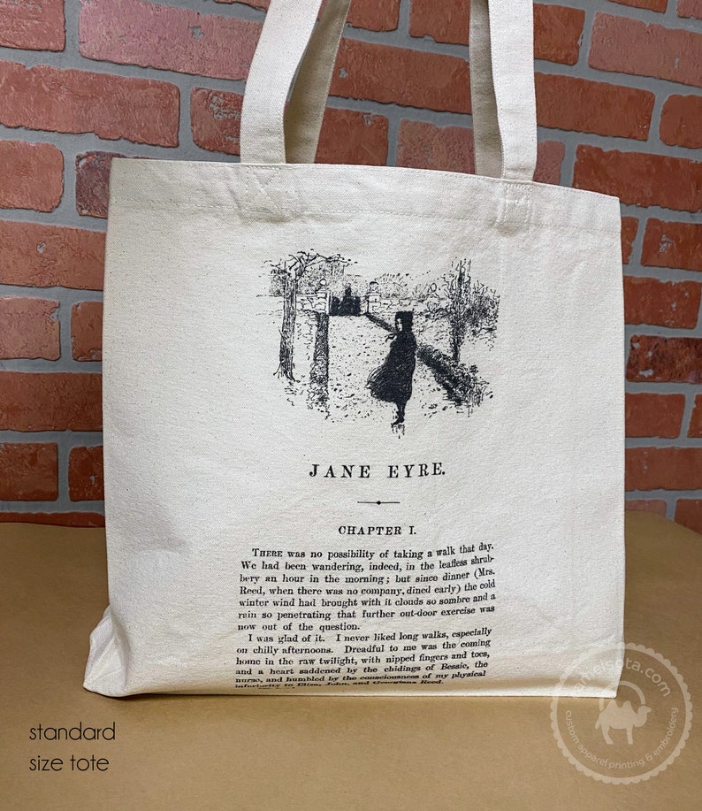 May include: A white canvas tote bag with black handles. The bag features a black and white illustration of a woman walking down a path, with trees on either side. The text "JANE EYRE" and "CHAPTER I." is printed on the bag.