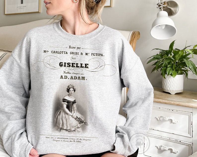 May include: Gray sweatshirt with a vintage-style print of a ballet poster. The poster features a black and white image of a ballerina in a white dress and a black bodice. The text on the poster reads "Danse par Mlle. CARLOTTA GRISI & M: PETIPA. GISELE Ballet. Compose par AD. ADAM."