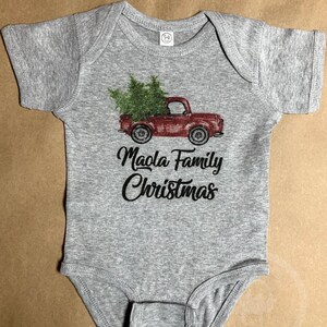 May include: A gray baby onesie with a red truck carrying green Christmas trees. The text "Maola Family Christmas" is printed in black script below the truck. The onesie has short sleeves and a round neckline.