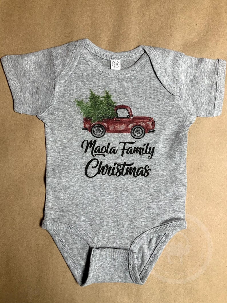 May include: A gray baby onesie with a red truck carrying green Christmas trees. The text "Maola Family Christmas" is printed in black script below the truck. The onesie has short sleeves and a round neckline.