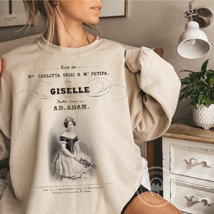 May include: A beige sweatshirt with a vintage-style print of a ballet poster. The poster features a black and white image of a ballerina with the text "Baner par M CARLOTTA GRISI & ME PETIPA. GISELLE Balla, compose par AD.ADAM."