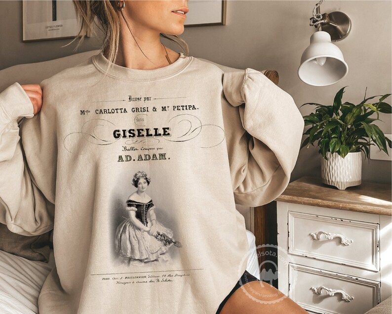 May include: A beige sweatshirt with a vintage-style print of a ballet poster. The poster features a black and white image of a ballerina with the text "Baner par M CARLOTTA GRISI & ME PETIPA. GISELLE Balla, compose par AD.ADAM."
