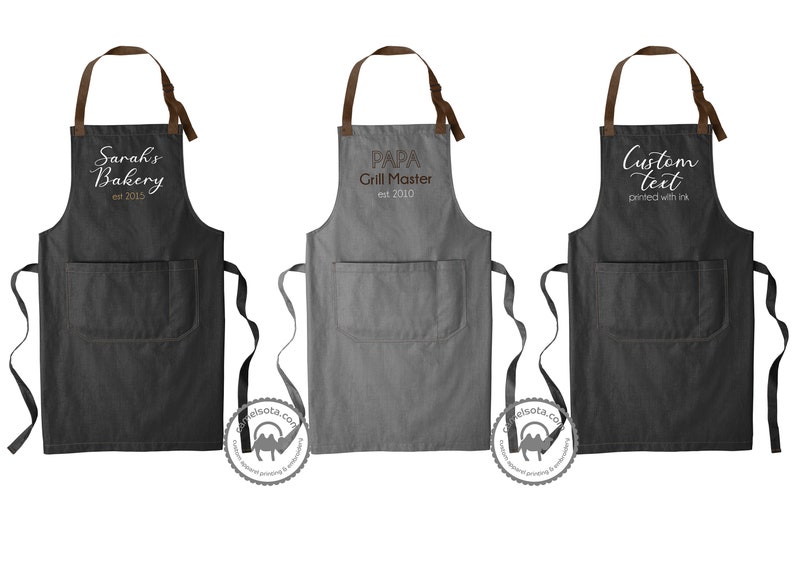 K&ouml;nnte beinhalten: Drei verschiedene Sch&uuml;rzen mit braunen Tr&auml;gern und einer Tasche. Die erste Sch&uuml;rze ist schwarz mit wei&szlig;em Text, der "Sarah's Bakery est 2015" lautet. Die zweite Sch&uuml;rze ist grau mit wei&szlig;em Text, der "PAPA Grill Master est 2010" lautet. Die dritte Sch&uuml;rze ist schwarz mit wei&szlig;em Text, der "Benutzerdefinierter Text mit Tinte gedruckt" lautet.