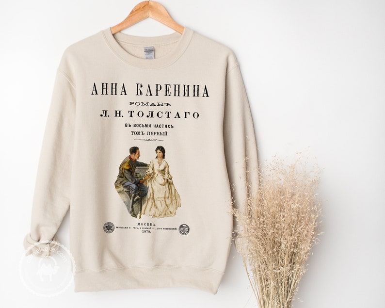 May include: A beige sweatshirt with a black print of the book cover for "Anna Karenina" by Leo Tolstoy. The cover features a vintage illustration of a man and woman in 19th-century clothing.