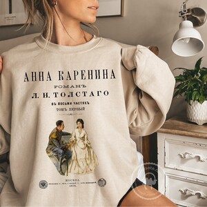May include: A beige sweatshirt with a vintage-style print of the book cover for "Anna Karenina" by Leo Tolstoy. The print features a black and white illustration of two people sitting on a bench.