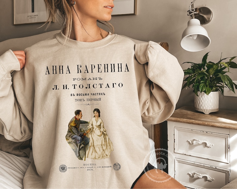 May include: A beige sweatshirt with a vintage-style print of the book cover for "Anna Karenina" by Leo Tolstoy. The print features a black and white illustration of two people sitting on a bench.