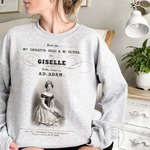 May include: Gray sweatshirt with a vintage-style print of a ballet poster. The poster features a black and white image of a ballerina in a white dress and a black bodice. The text on the poster reads "Danse par Mlle. CARLOTTA GRISI & M: PETIPA. GISELE Ballet. Compose par AD. ADAM."