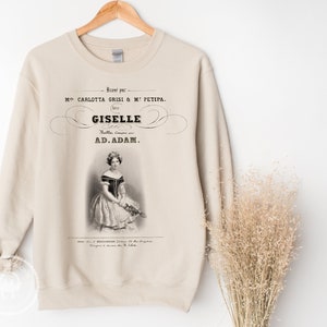 May include: A beige sweatshirt with a vintage-style print of a ballerina in a white dress. The print includes the text "Giselle" and "Ad. Adam."