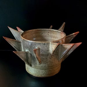 May include: A ceramic bowl with a speckled beige and brown glaze, featuring multiple triangular, spike-like protrusions extending outward from the sides. The vessel has a wide, open circular rim and a solid, flat base.