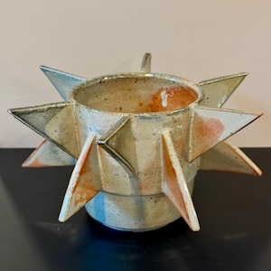Peut inclure: Un pot en céramique avec un corps cylindrique et huit pointes triangulaires saillantes sur les côtés. La surface présente un émail crème moucheté avec des touches de tons orange et gris, créant un aspect texturé et irrégulier.