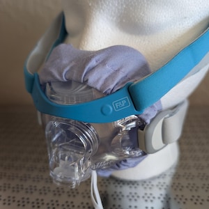 May include: A clear plastic CPAP mask with a light blue silicone frame featuring the F&P logo. The mask includes a soft, gathered lavender fabric cover over the nose bridge area and light blue elastic headgear straps with grey plastic connectors.
