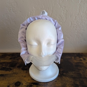 Peut inclure: Un bonnet en satin violet clair sur une tête de mannequin blanche. Le bonnet est conçu pour couvrir la tête et protéger les cheveux. Le mannequin est sur une surface en bois foncé.