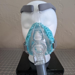 Könnte beinhalten: Eine transparente und blaue CPAP-Maske mit grauen Riemen und einem türkisfarbenen Kissen wird auf einem weißen Schaufensterkopf gezeigt. Auf der Maske steht "F&P Simplus".