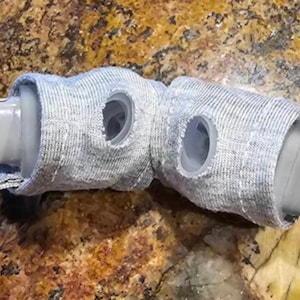Peut inclure: Deux pièces en plastique grises recouvertes d'une gaine en tissu gris. Le tissu comporte des trous découpés pour exposer les pièces en plastique.