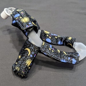 May include: A clear plastic CPAP mask with black fabric covers featuring a celestial print of stars, moons, and suns in yellow, blue, and pink.