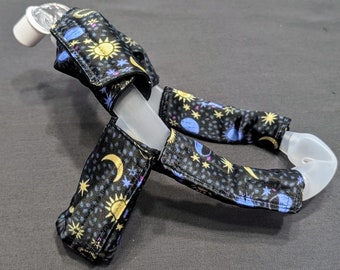 Può includere: Una maschera CPAP in plastica trasparente con rivestimenti in tessuto nero che presentano una stampa celeste di stelle, lune e soli in giallo, blu e rosa.