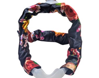 Può includere: Una copertura per maschera CPAP in raso nero con un vivace motivo floreale in rosso, rosa e giallo. La copertura è avvolta attorno a un telaio in plastica trasparente curvo e a un pezzo superiore in plastica trasparente.