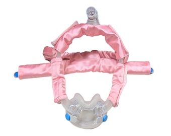 Può includere: Una maschera CPAP con una copertura facciale in plastica trasparente e un'imbottitura in tessuto di raso rosa. La maschera ha dettagli blu e un connettore per tubi in plastica trasparente. L'imbottitura è progettata per il comfort.