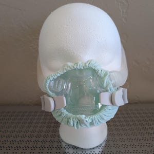 May include: A clear plastic face mask with a pale blue fabric trim and adjustable grey straps. The mask is mounted on a white foam head. The mask is designed to cover the nose and mouth.