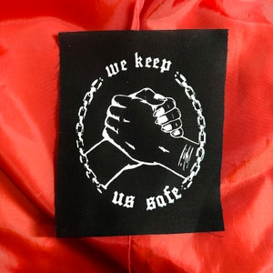 May include: Black square patch with white text and graphic. The text reads "we keep us safe" in a gothic font. The graphic depicts two hands clasped in a handshake, surrounded by a chain. The patch is on a red background.