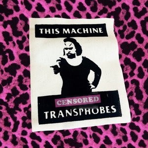May include: A white canvas patch with black text and a black silhouette of a person pointing. The text reads "THIS MACHINE" above the image and "CENSORED" and "TRANSPHOBES" below. The background is a pink leopard print.