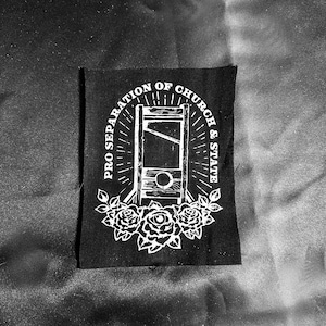 May include: Black patch with white graphic of a guillotine, surrounded by text reading "PRO SEPARATION OF CHURCH & STATE" and roses. The patch is rectangular and likely made of fabric.