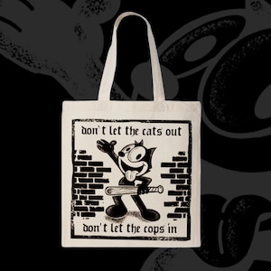 May include: Off-white tote bag featuring a black and white cartoon cat holding a baseball bat, with the text "don't let the cats out" and "don't let the cops in". The cat is standing in front of a brick wall design.