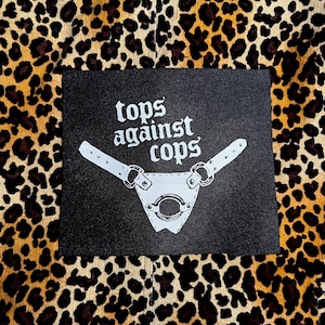 May include: A black patch with the words "tops against cops" in white, stylized lettering. Below the text is a white leather-like design with a circular opening and straps. The background is a leopard print pattern.