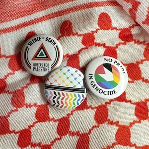 May include: Three circular badges with political statements. One says "SILENCE = DEATH" with a triangle and "QUEERS FOR PALESTINE." Another says "NO PRIDE IN GENOCIDE" with a rainbow design. The third has a rainbow pattern.