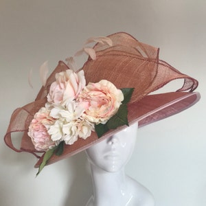 May include: A blush pink wide-brimmed hat with a large pink and white flower arrangement. The hat is made of a delicate, woven material and has a soft, feminine look.
