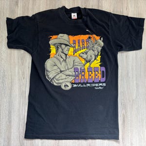 May include: Black t-shirt with a graphic of a cowboy riding a bull. The graphic is in yellow, orange, purple, and white. The text on the shirt reads "Rare Breed Bullriders".