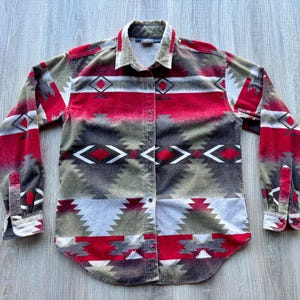 Puede incluir: Una camisa abotonada con un estampado de inspiración suroeste. La camisa presenta una mezcla de diseños geométricos rojos, grises, blancos y verde oliva. Tiene mangas largas y cuello. La camisa está hecha de una tela suave y texturizada.