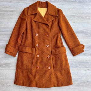 May include: A double-breasted coat in a textured burnt orange wool-like fabric featuring a wide notched collar, long sleeves with buttoned cuffs, and two large front patch pockets. The coat is secured with eight round brown buttons.