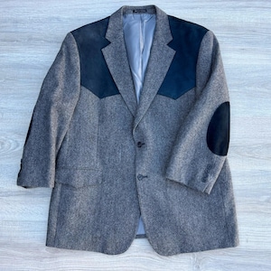 Puede incluir: Un blazer gris con parches de ante negro en los hombros y los codos. El blazer tiene cierre de dos botones y forro azul claro. Las mangas tienen botones en los puños. El blazer está hecho de una tela texturizada.