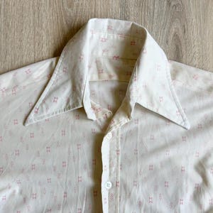 May include: A cream-colored button-down shirt featuring a repeating pattern of small, light pink geometric shapes. The shirt has a pointed collar, a front button placket with visible white buttons, and long sleeves with a textured fabric finish.