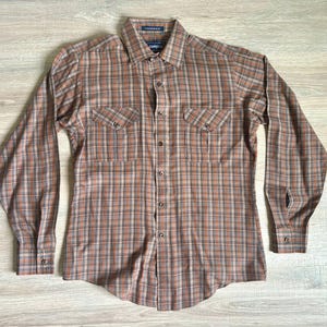 Puede incluir: Una camisa de manga larga a cuadros con un patrón de cuadros marrones y naranjas. La camisa tiene un cuello abotonado y dos bolsillos en el pecho.