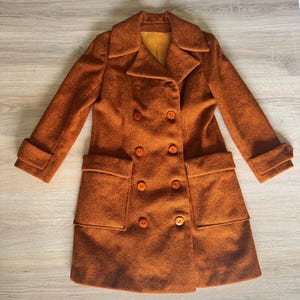 Puede incluir: Un abrigo vintage de doble botonadura en un cálido tono naranja. El abrigo presenta botones grandes y redondos, dos bolsillos delanteros y un cuello clásico. Las mangas están dobladas y el abrigo tiene un corte entallado. El forro interior es de color amarillo brillante.