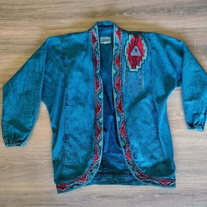 May include: A turquoise denim jacket with an open front and long sleeves. The jacket features a colorful embroidered design on the lapels and upper back, with red, turquoise, and purple accents. The jacket is laid flat on a wooden surface.