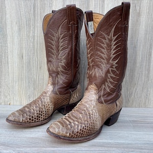 May include: A pair of brown leather cowboy boots with snake skin vamps. The boots have a pointed toe and decorative stitching on the shaft. The boots are set against a light wood grain background.