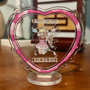 May include: A clear acrylic heart-shaped stand with a pink camouflage border. The stand holds a cartoon character in a pink dress with angel wings. The base is a clear, circular shape.